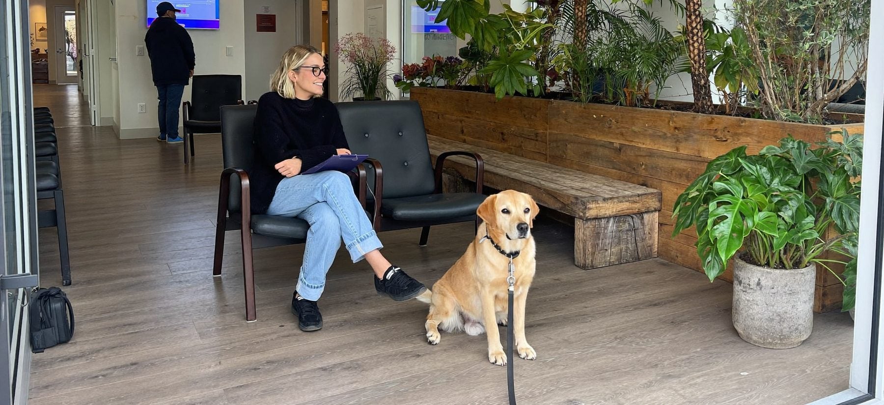 A woman waits in a bright vet clinic waiting room with her golden Labrador sitting beside her.